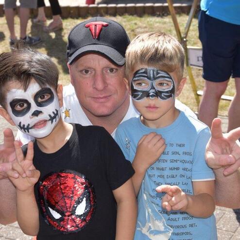 Mann umarmt zwei Kinder mit Gesichtsbemalung, einer mit Totenkopf-Muster, der andere als Katze, alle drei zeigen Peace-Zeichen.