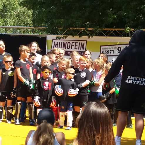 Gruppierung von Kindern in Sportkleidung auf einer B&uuml;hne, mit einem Trainer, vor einem Banner im Hintergrund.