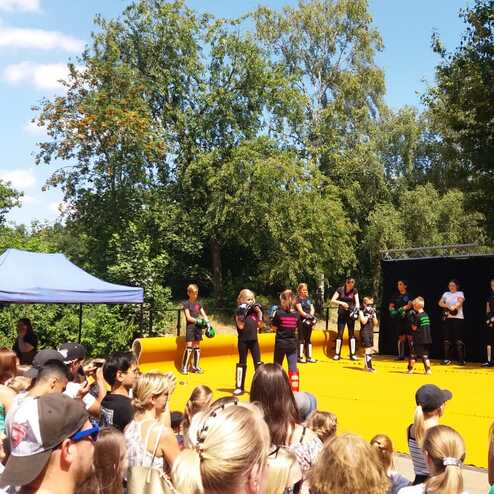 Gruppe von Menschen auf einer Veranstaltung im Freien, umgeben von Zuschauern unter blauem Himmel und B&auml;umen.