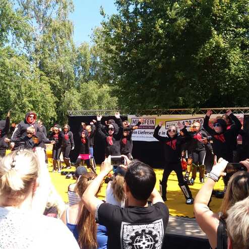 Tanzgruppe in schwarz-roten Outfits f&uuml;hrt eine Auff&uuml;hrung auf einer gelben B&uuml;hne vor einem Publikum im Freien auf.