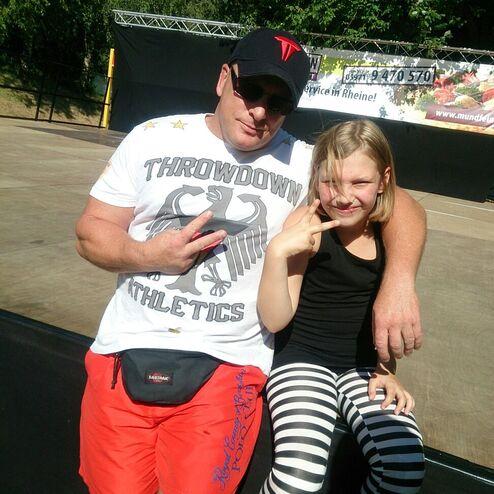 Mann mit Sonnenbrille und Kind in gestreifter Hose posieren l&auml;chelnd auf einer B&uuml;hne im Freien.