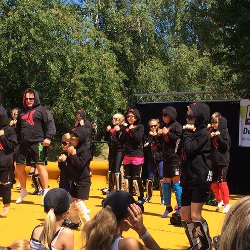 Gruppenvorstellung von Kindern und Erwachsenen in Trainingsanz&uuml;gen auf einer Au&szlig;enb&uuml;hne mit gr&uuml;nen B&auml;umen im Hintergrund.