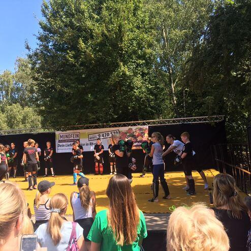 Menschen stehen auf einer B&uuml;hne und tanzen, w&auml;hrend eine Gruppe Zuschauer im Vordergrund zusieht.