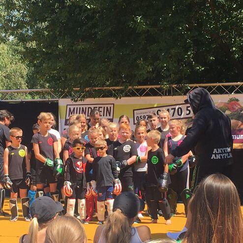 Gruppe von Kindern in Sportkleidung mit Boxhandschuhen, die auf einer Wettkampf-B&uuml;hne stehen und einem Trainer zuh&ouml;ren.