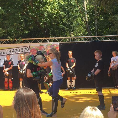 Kinder trainieren in Schutzausr&uuml;stung beim Kampfsport-Event auf einer gelben Matte unter freiem Himmel.