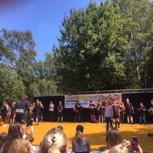 Gruppentraining auf einer gelben Fl&auml;che im Freien, umgeben von Zuschauern und B&auml;umen.