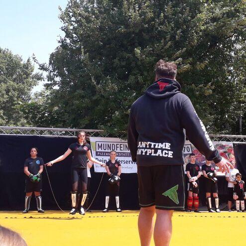 Gruppen von Kindern und Jugendlichen in Sportkleidung stehen auf einer gelben Fl&auml;che, w&auml;hrend ein Trainer Anweisungen gibt.