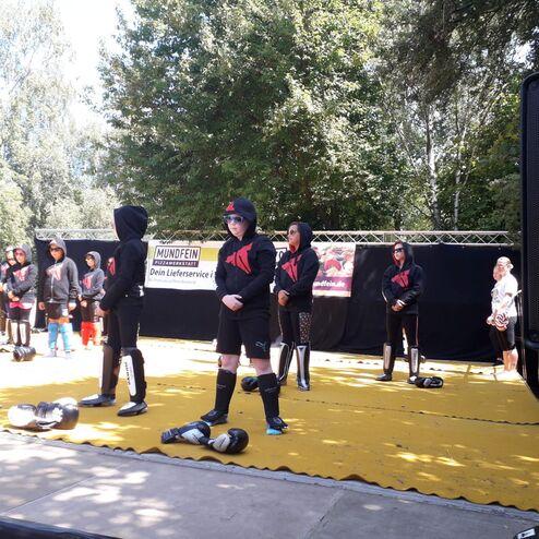 Gruppe von Personen in schwarzer Aktivkleidung steht auf einer gelben Fl&auml;che, umgeben von B&auml;umen und Zuschauer.