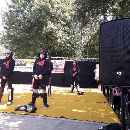 Gruppe von Personen in Sportkleidung steht auf einer gelben Matte; Lautsprecher im Vordergrund, B&auml;ume im Hintergrund.