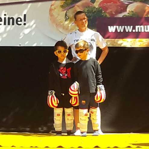 Zwei Kinder mit Handschuhen und Sonnenbrillen stehen auf einer gelben Matte, im Hintergrund ein Trainer und ein Werbebanner.