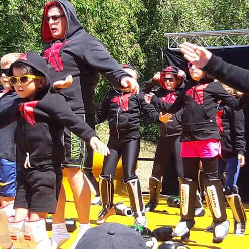 Gruppe von Kindern und Erwachsenen in schwarzen Kapuzenpullovern mit Sonnenbrillen beim Tanzen auf einer B&uuml;hne im Freien.