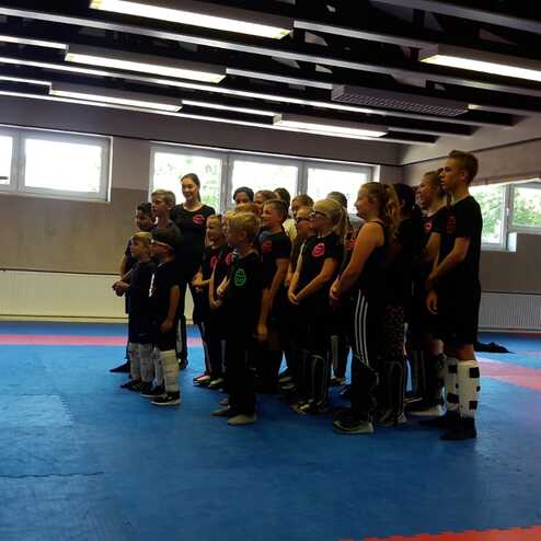 Gruppe von Kindern in Sportkleidung steht auf einer blauen Matte in einem Trainingsraum, w&auml;hrend ein Trainer Anweisungen gibt.