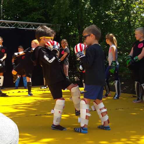 Zwei Kinder im Trainingsanzug &uuml;ben Boxtechniken auf einer gelben Trainingsfl&auml;che, umgeben von anderen jungen K&auml;mpfern.