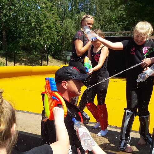 Kinder spritzen mit Wasserpistolen auf einen Erwachsenen, der auf einer gelben Fl&auml;che steht, w&auml;hrend eine sommerliche Aktivit&auml;t stattfindet.