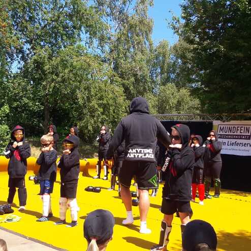 Kinder in Sportkleidung stehen auf einer gelben Plattform und zeigen Kampfsport&uuml;bungen unter Anleitung eines Trainers.