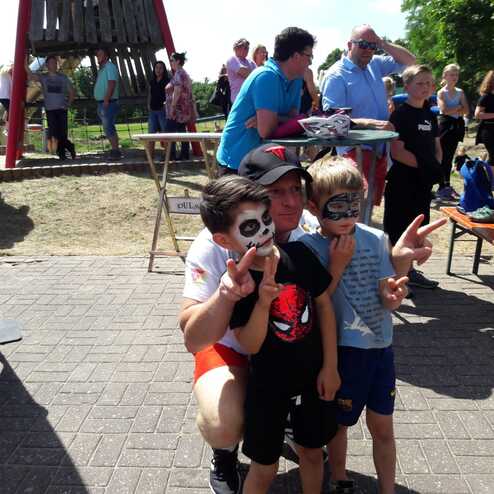 Drei Kinder posieren im Freien, eines tr&auml;gt Gesichtsfarbe, das andere einen Spiderman-Anzug, im Hintergrund Menschen und St&auml;nde.