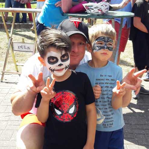 Drei Personen posieren l&auml;chelnd im Freien, zwei Kinder mit Gesichtsfarbe und ein Erwachsener hinten.