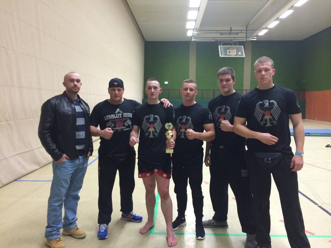 Sechs Personen in Sportbekleidung stehen in einer Halle, einer h&auml;lt einen Pokal und alle zeigen den SiegervSymbol.