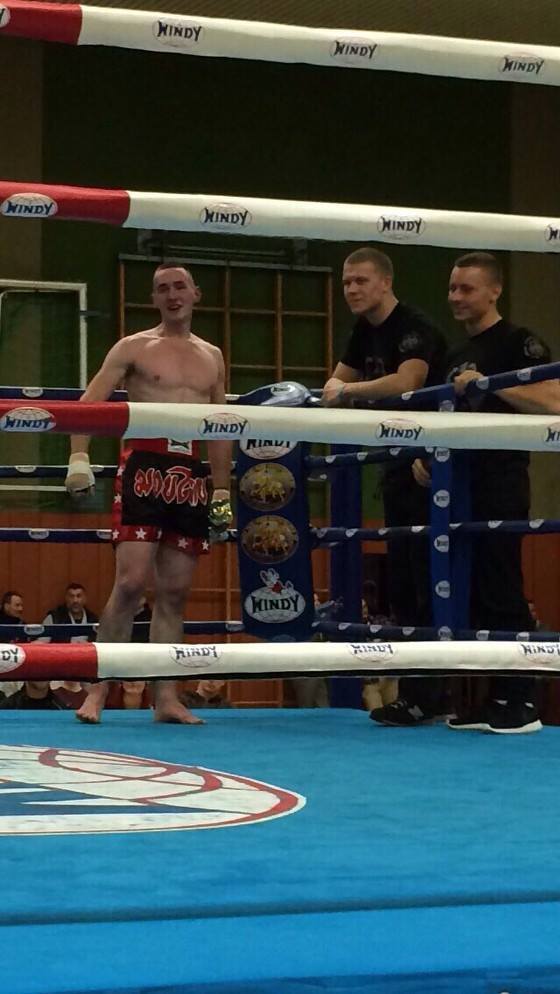 Ein unbedeckter K&auml;mpfer in Boxshorts steht im Ring, umgeben von zwei Trainern, w&auml;hrend Zuschauer im Hintergrund warten.