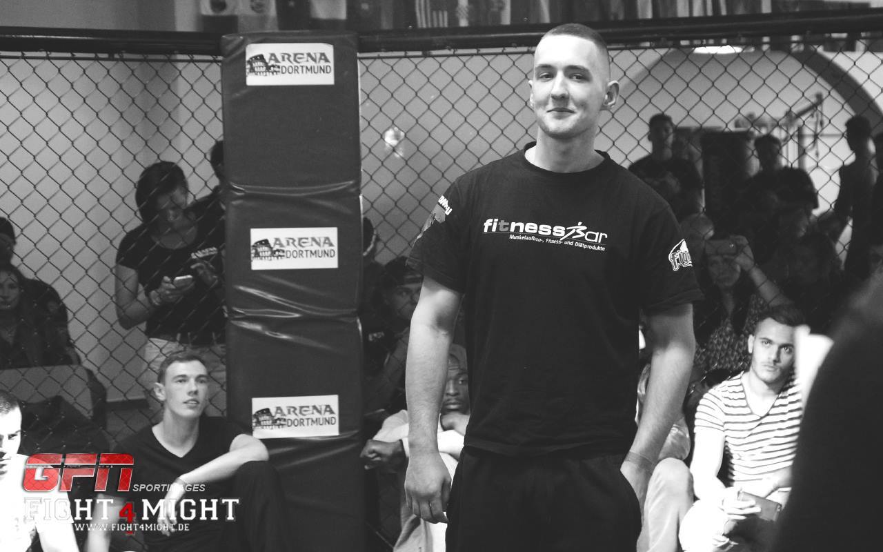 Ein Mann in schwarzem T-Shirt steht l&auml;chelnd vor einem K&auml;fig mit Zuschauern im Hintergrund.