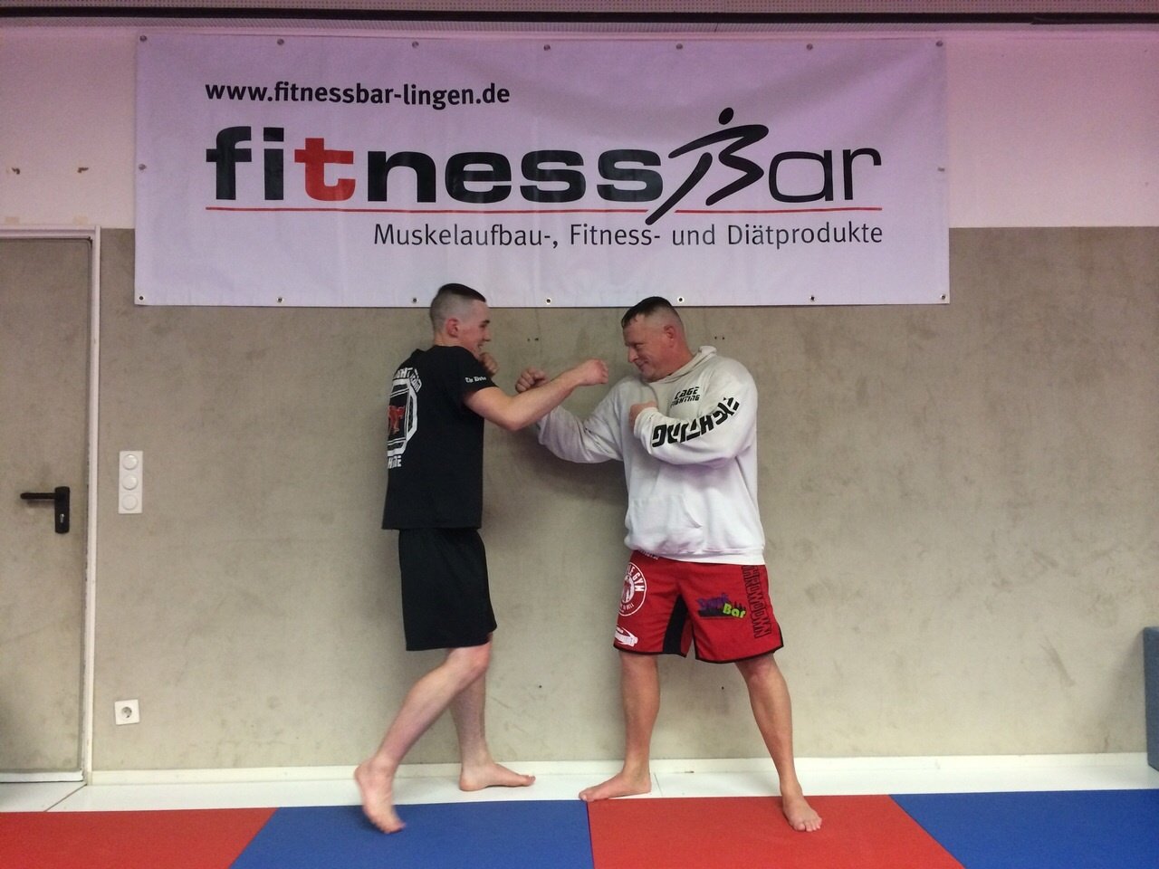 Zwei M&auml;nner im Fitnessstudio &uuml;ben einen Griff in einer Kampfsport-Trainingseinheit vor einem Fitnessbanner.