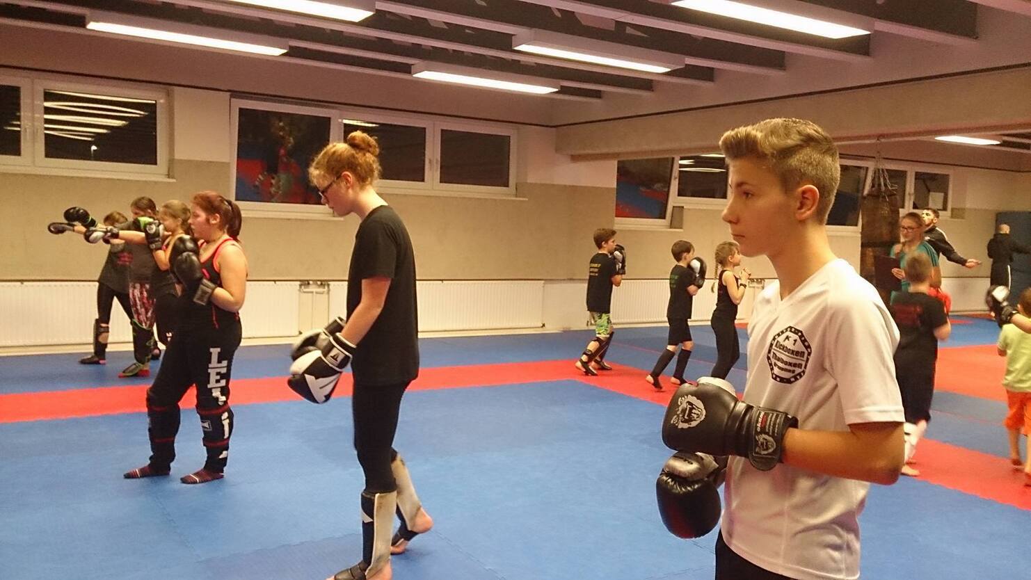 Trainingseinheit im Kampfsport mit Kindern und Jugendlichen in einem Sportraum, Boxhandschuhe und &Uuml;bungshaltung sichtbar.