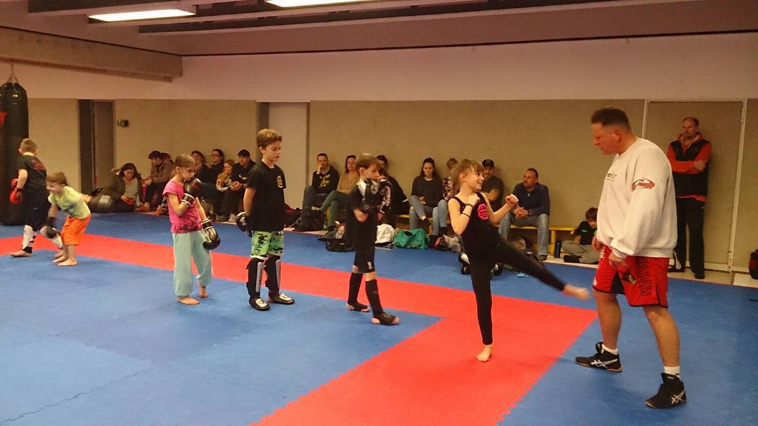 Kinder &uuml;ben Kampfsporttechniken in einem Training mit einem Trainer in einem Sportstudio. Zuschauer sitzen im Hintergrund.