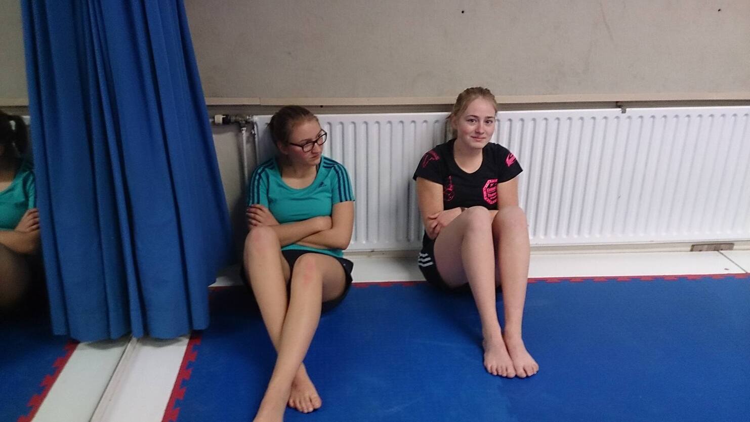 Zwei M&auml;dchen sitzen mit angezogenen Beinen an einer Wand auf einer blauen Matte in einem Trainingsraum.