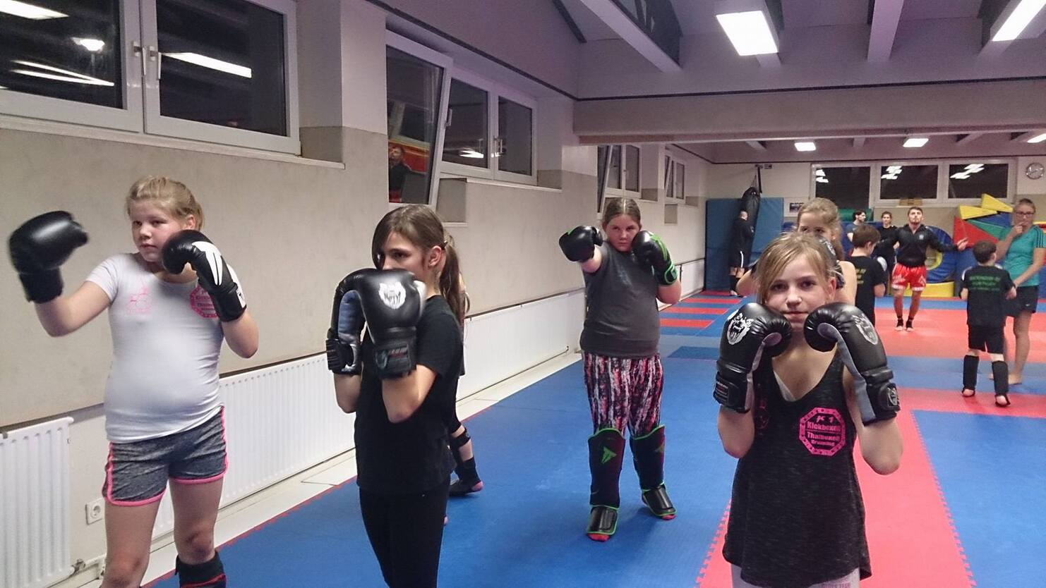 Aktive M&auml;dchen trainieren im Karate-Anzug mit Boxhandschuhen in einem Sportraum auf blauen Matten.