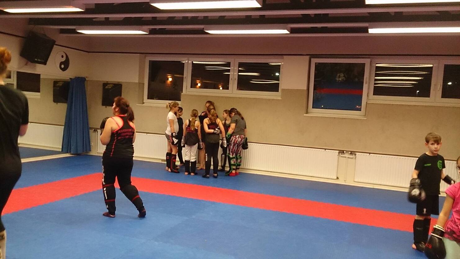 Gruppentraining im Kampfsportraum mit mehreren Personen in Schutzkleidung auf blauer und roter Matte.