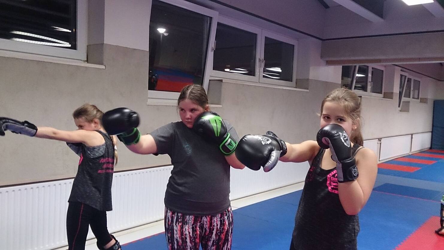 Drei M&auml;dchen in Sportbekleidung &uuml;ben Boxen in einem Trainingsraum, zwei davon tragen Boxhandschuhe.