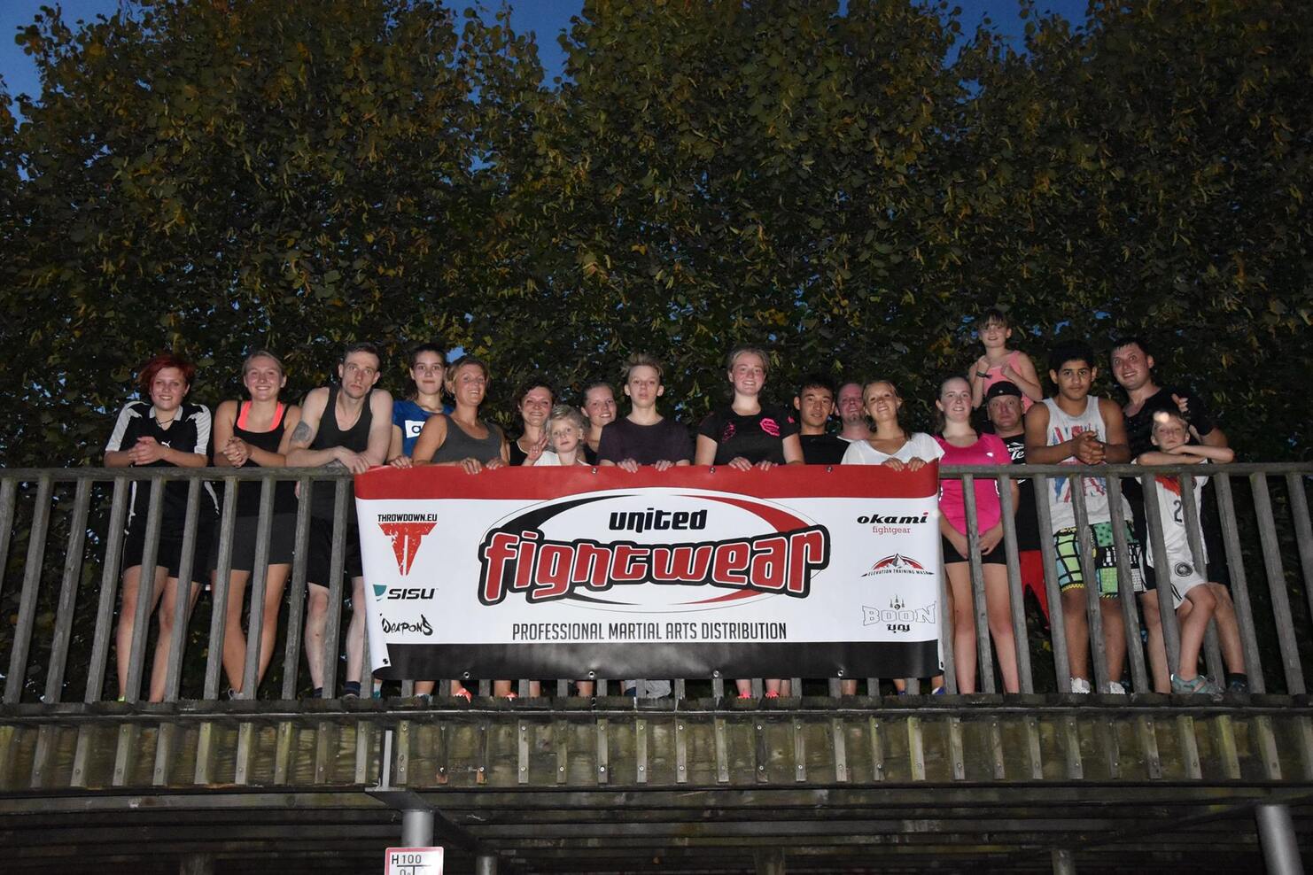 Gruppenfoto von 20 Personen auf einer Holzplattform, ein Banner mit der Aufschrift &bdquo;united fightwear&ldquo; sichtbar.