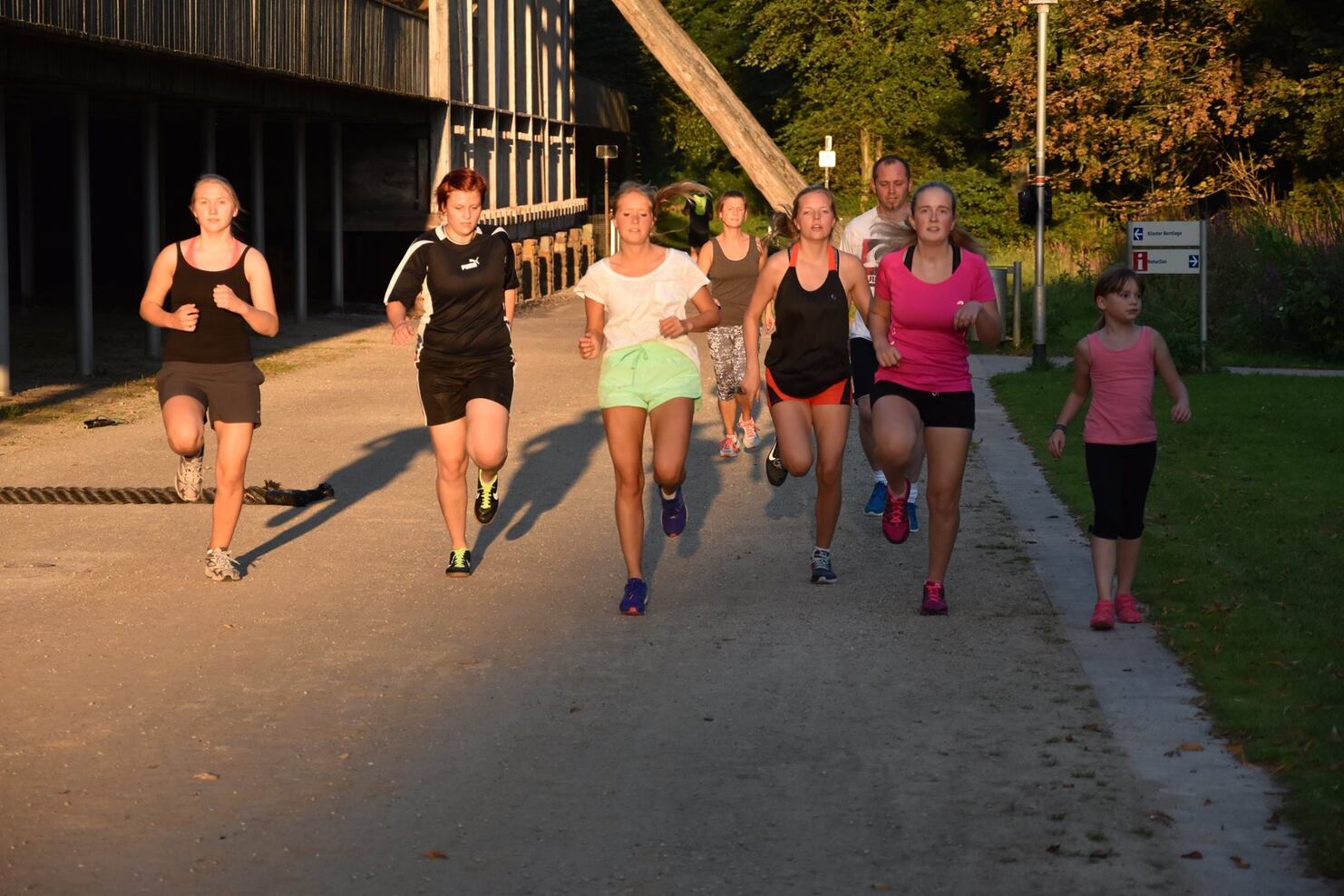 Gruppe von sieben Personen joggt entlang eines Wegs, umgeben von B&auml;umen und einem Geb&auml;ude im Hintergrund.