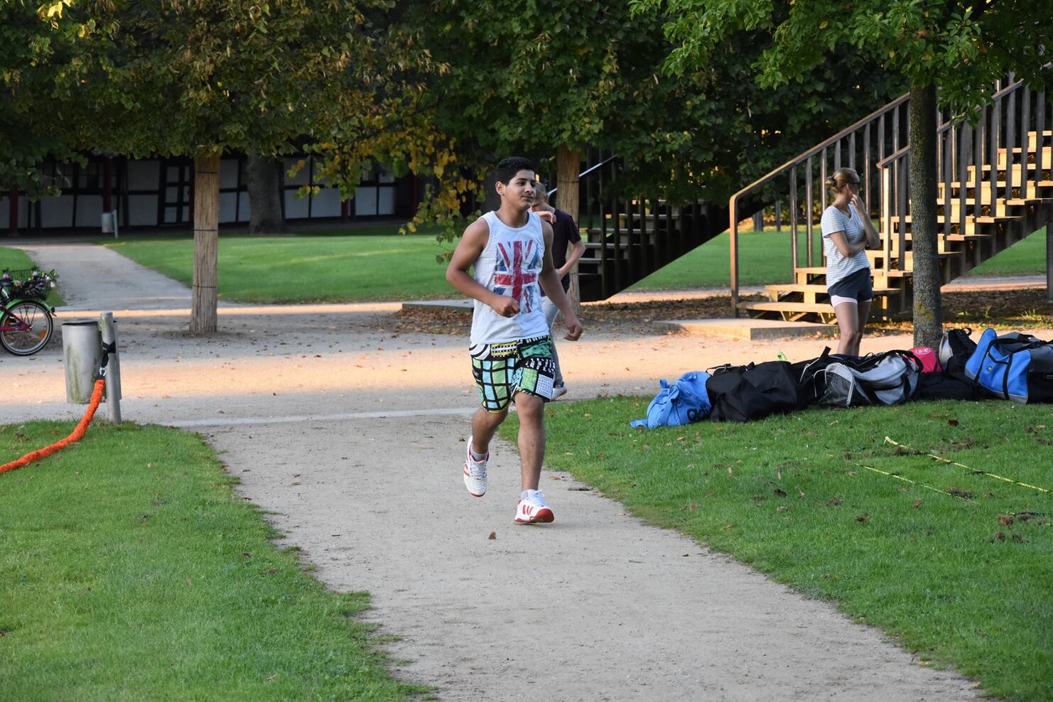Ein junger Mann l&auml;uft joggend auf einem Weg, w&auml;hrend im Hintergrund eine Frau mit Taschen besch&auml;ftigt ist.