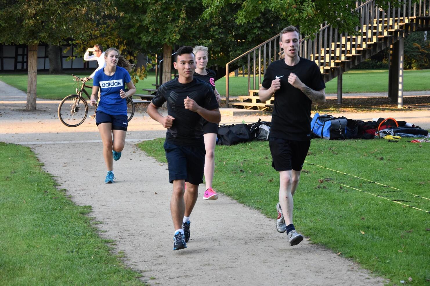 Vier Personen joggen auf einem Fu&szlig;weg im Park, w&auml;hrend eine Frau im Hintergrund Fahrrad f&auml;hrt.