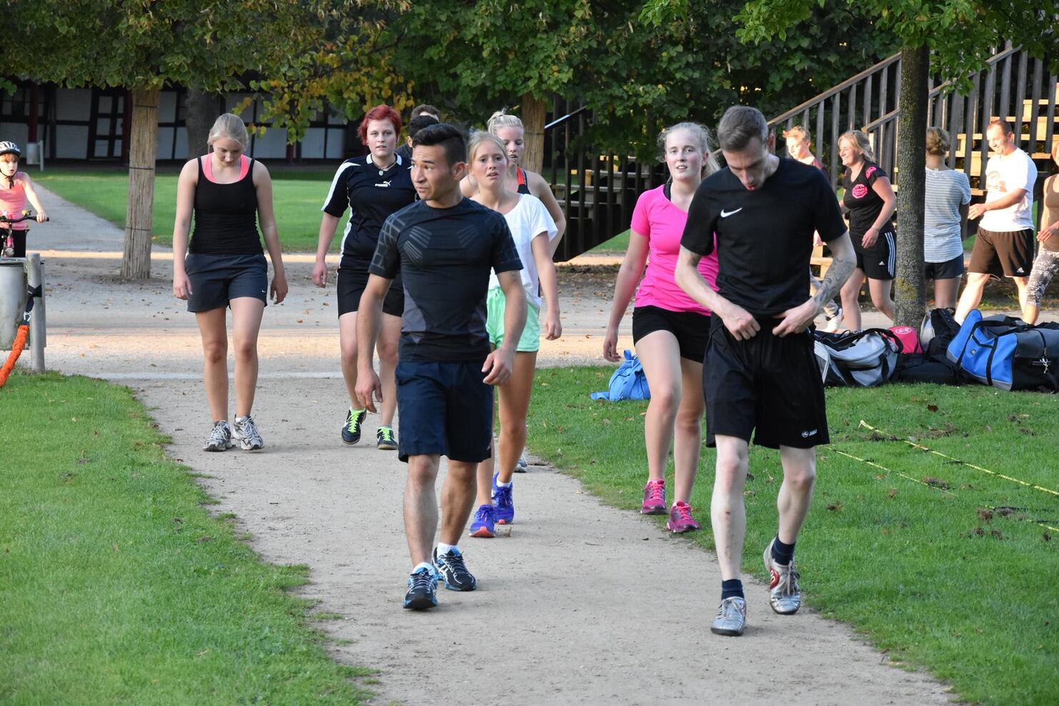 Gruppe von Sportlern l&auml;uft auf einem Schotterweg, umgeben von B&auml;umen und Sporttaschen im Hintergrund.