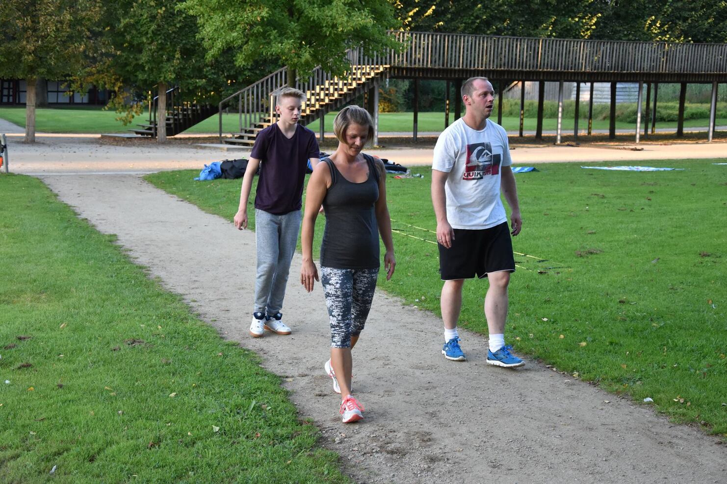 Drei Personen gehen auf einem schmalen Weg in einem Park, umgeben von Gras und B&auml;umen.