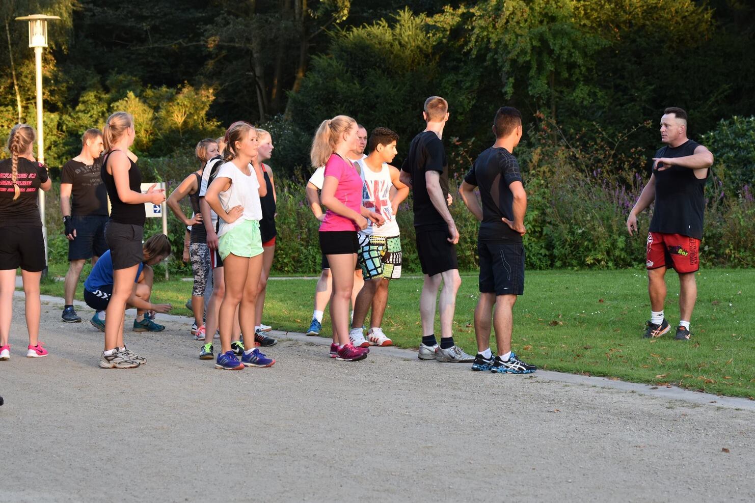 Gruppe von Jugendlichen in Sportkleidung h&ouml;rt einem Trainer im Freien aufmerksam zu. Hintergrund mit B&auml;umen und Wiese.