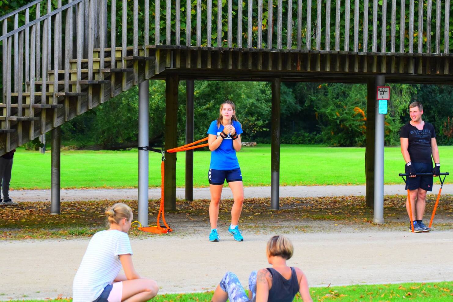 F&uuml;nf Personen trainieren im Freien: eine Frau nutzt ein Widerstandsband, w&auml;hrend andere entspannt sitzen oder stehen.