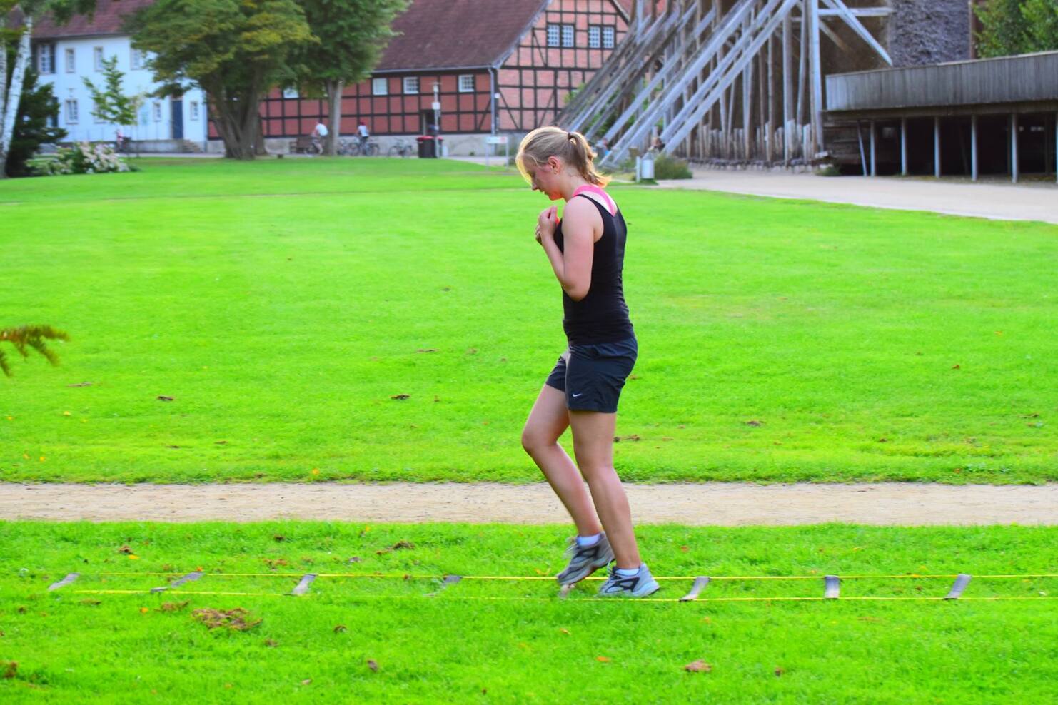 Junge Frau trainiert auf einem Schwebebalken im Freien, umgeben von gr&uuml;nem Rasen und historischen Geb&auml;uden im Hintergrund.