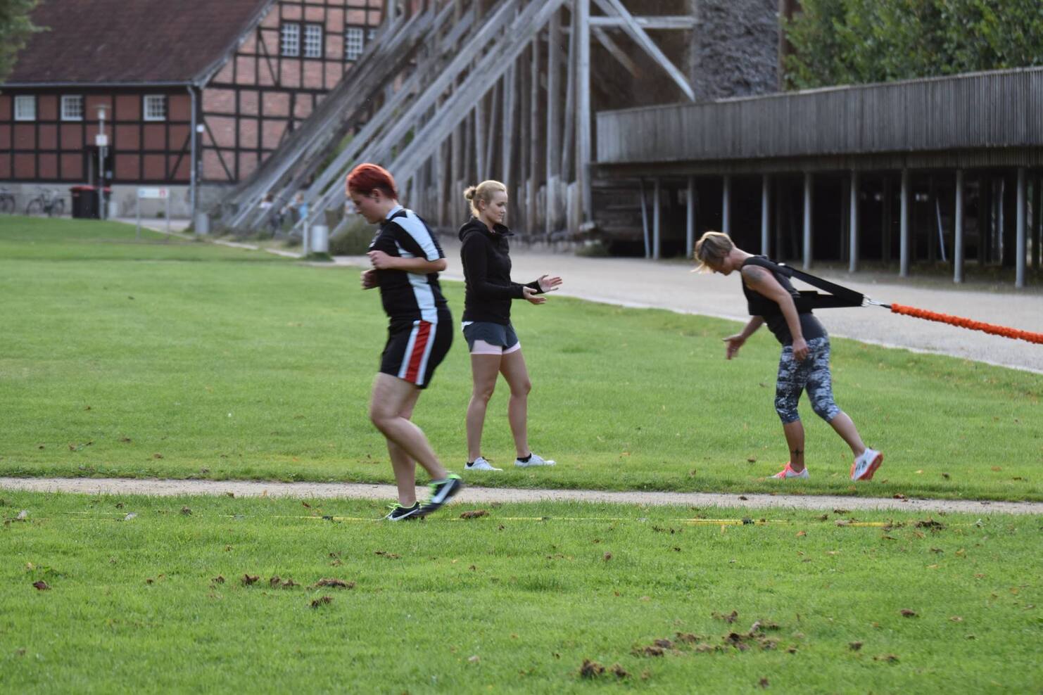 Drei Personen &uuml;ben eine Sportart auf einer Wiese, w&auml;hrend ein Holzgeb&auml;ude im Hintergrund sichtbar ist.