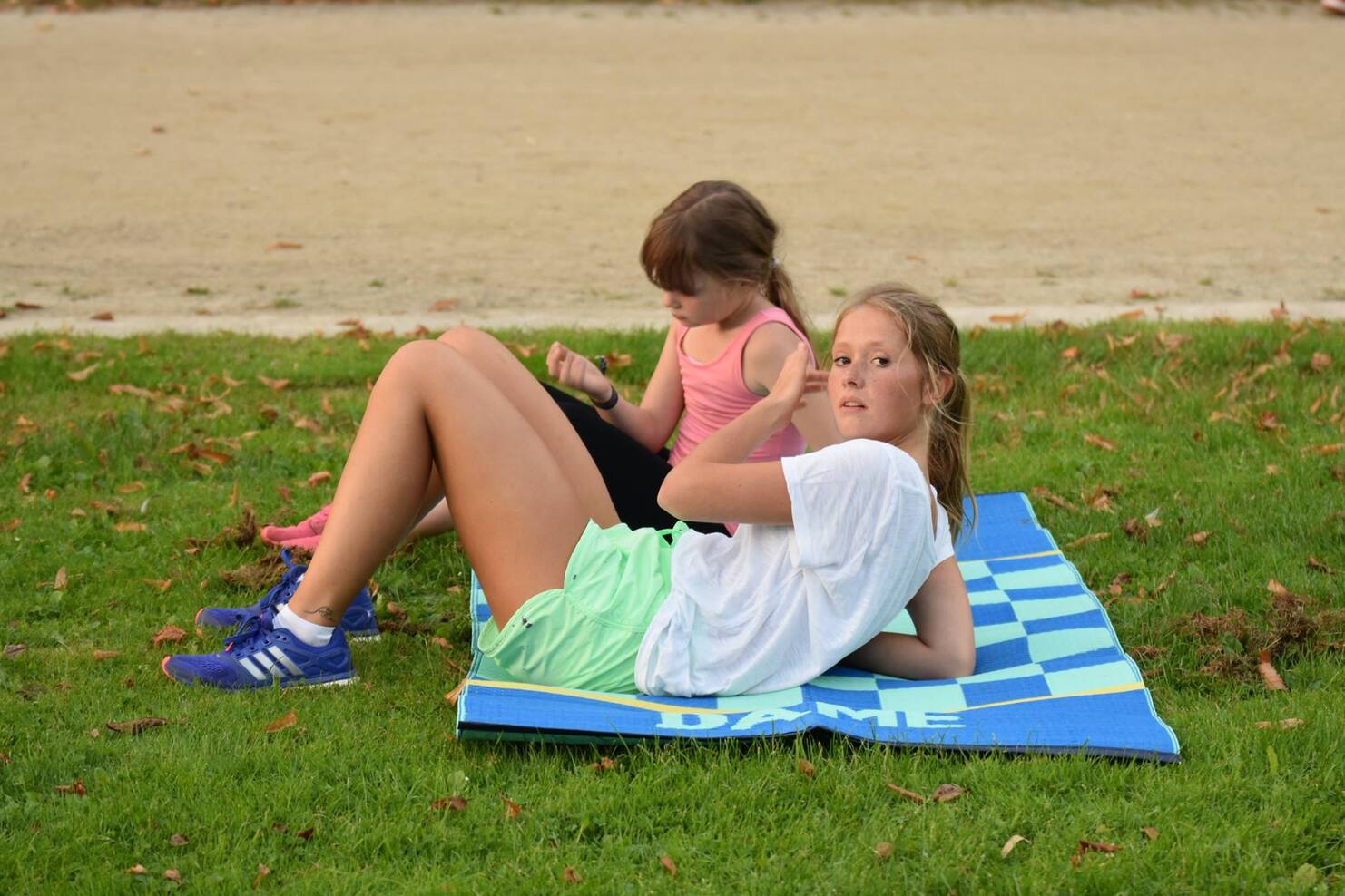 Zwei M&auml;dchen &uuml;ben im Freien; eines macht Sit-ups auf einer Matte, das andere sitzt daneben.