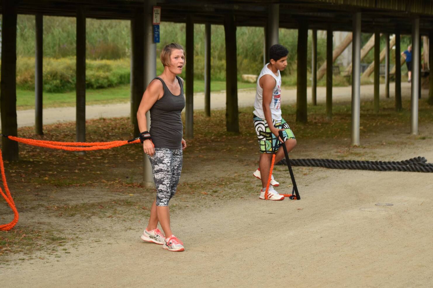 Zwei Personen trainieren im Freien: Eine Frau mit Gummiseil und ein Mann mit einem Stock in sportlicher Kleidung.