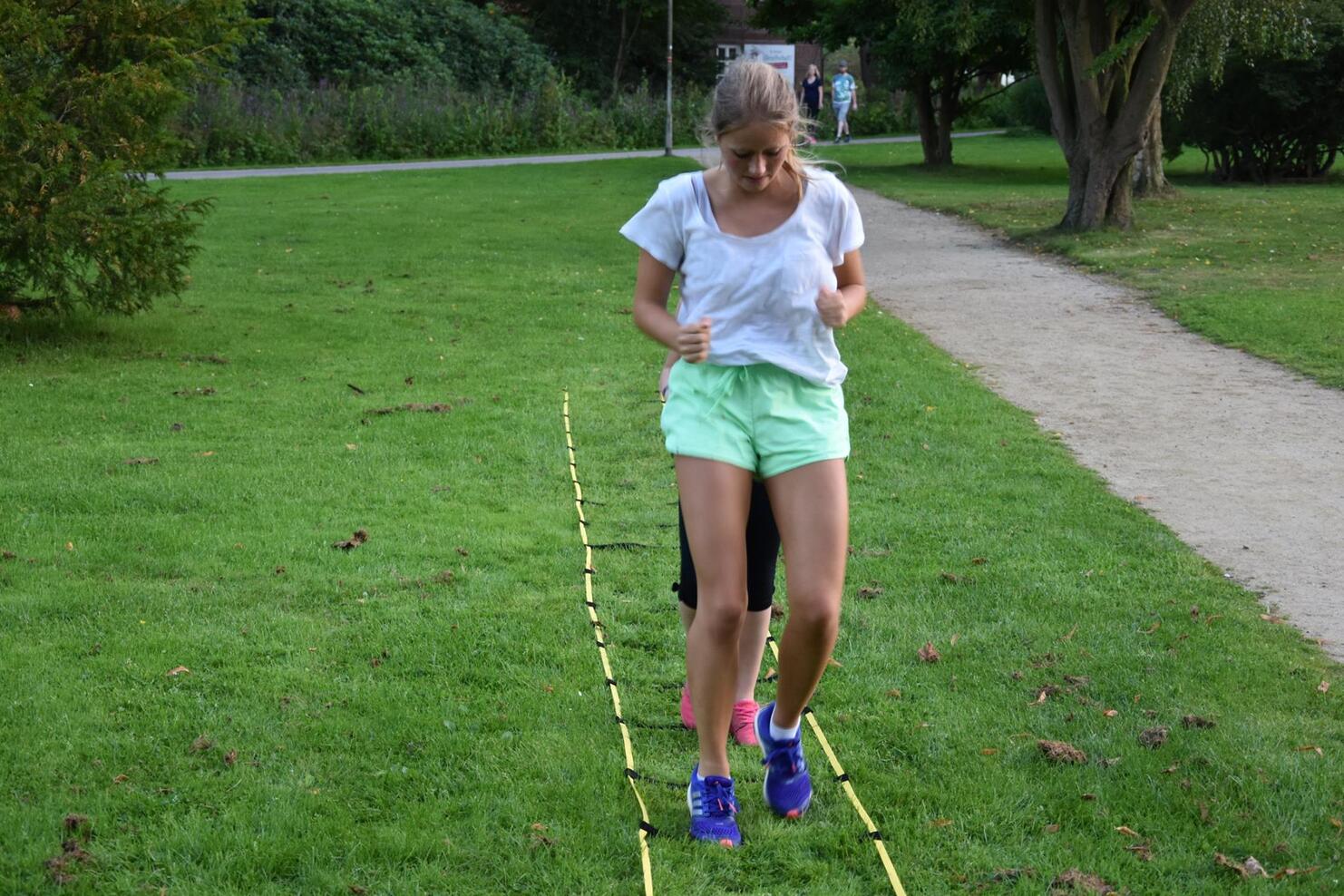 Junge Frau mit kurzen Hosen springt &uuml;ber Bodenmarkierungen in einem Park. Im Hintergrund ist ein Spazierweg zu sehen.