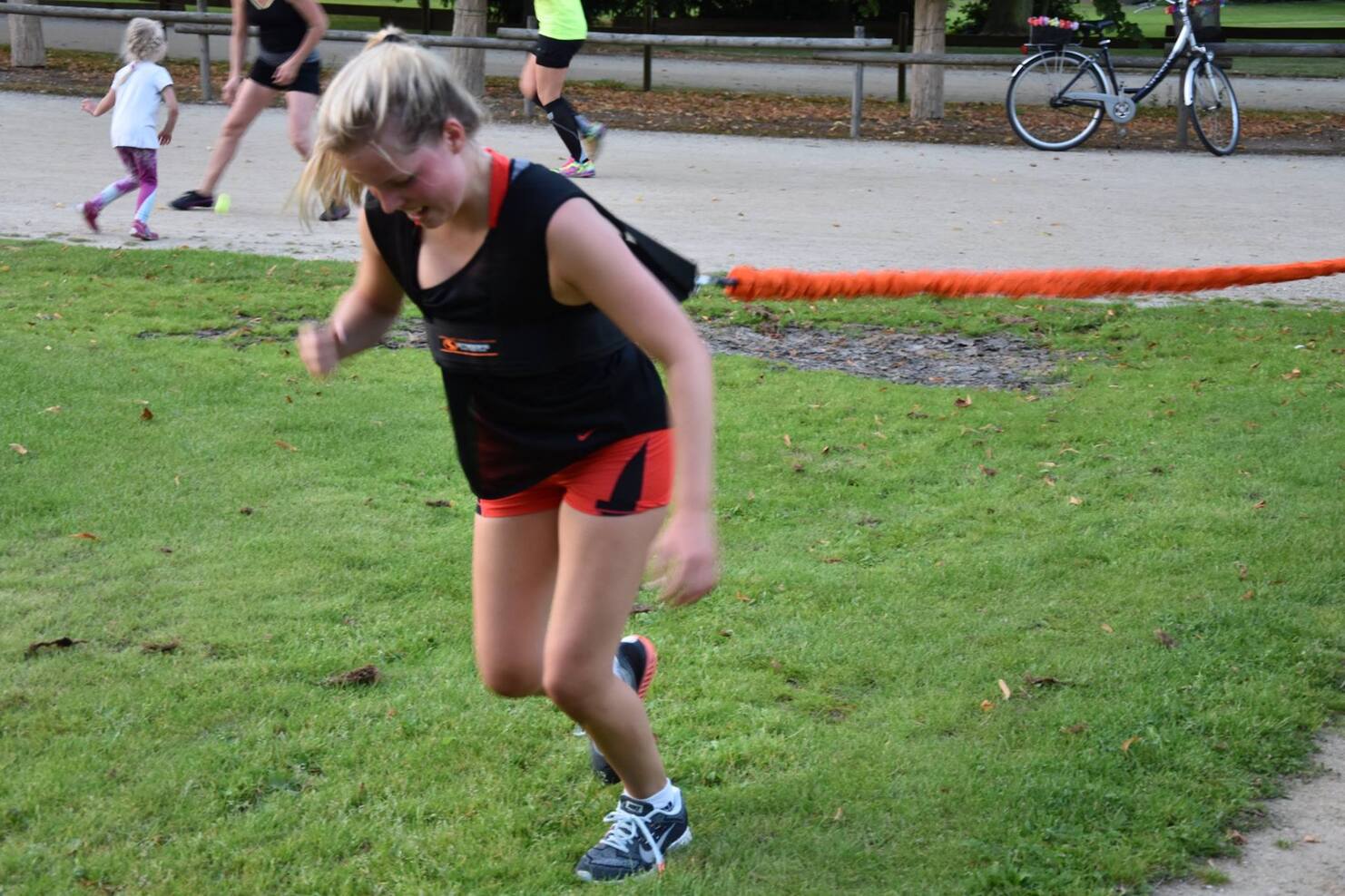 Eine Frau in Sportbekleidung sprintet auf einer Rasenfl&auml;che, w&auml;hrend andere im Hintergrund joggen.
