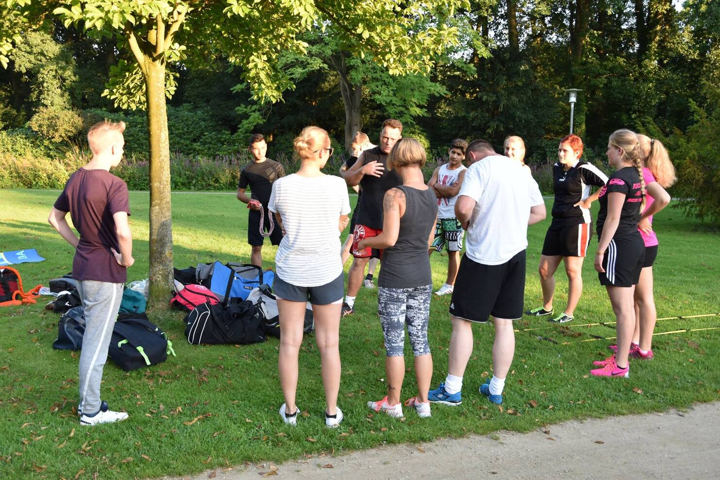 Gruppe von Menschen in Sportkleidung versammelt sich im Freien um einen Trainer unter einem Baum.