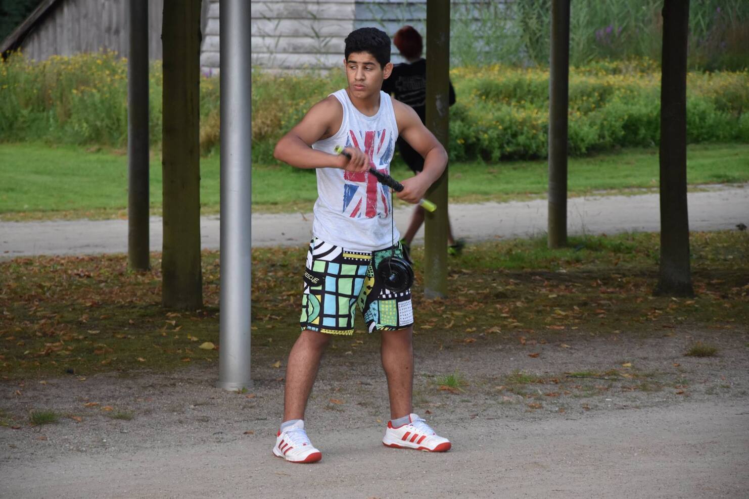 Junger Mann in sportlicher Kleidung mit Schl&auml;ger steht auf einem Weg in einem Park mit Blumenbeeten im Hintergrund.