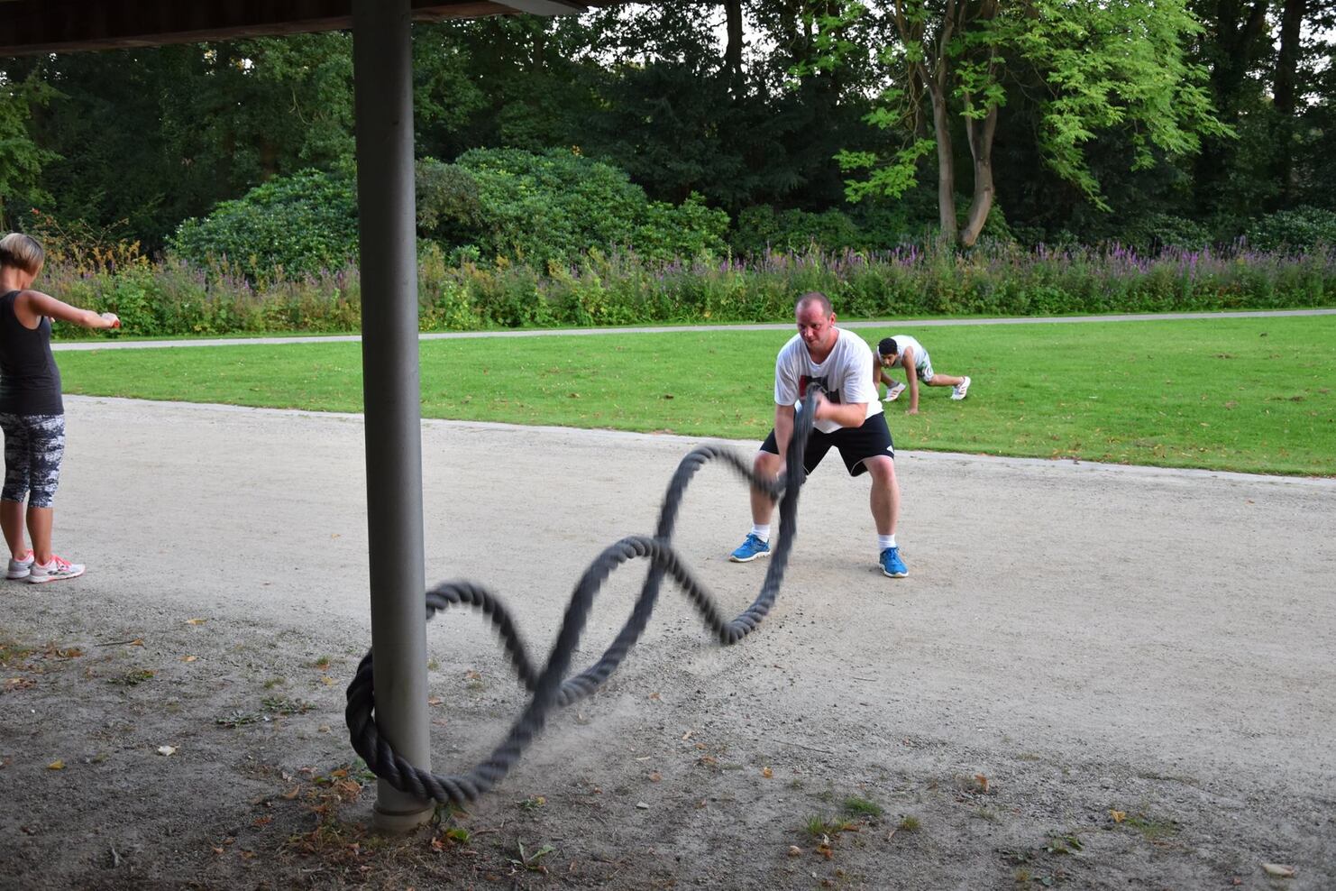 Mann trainiert mit einem Battle Rope auf einem Schotterweg, w&auml;hrend zwei weitere Personen im Hintergrund aktiv sind.