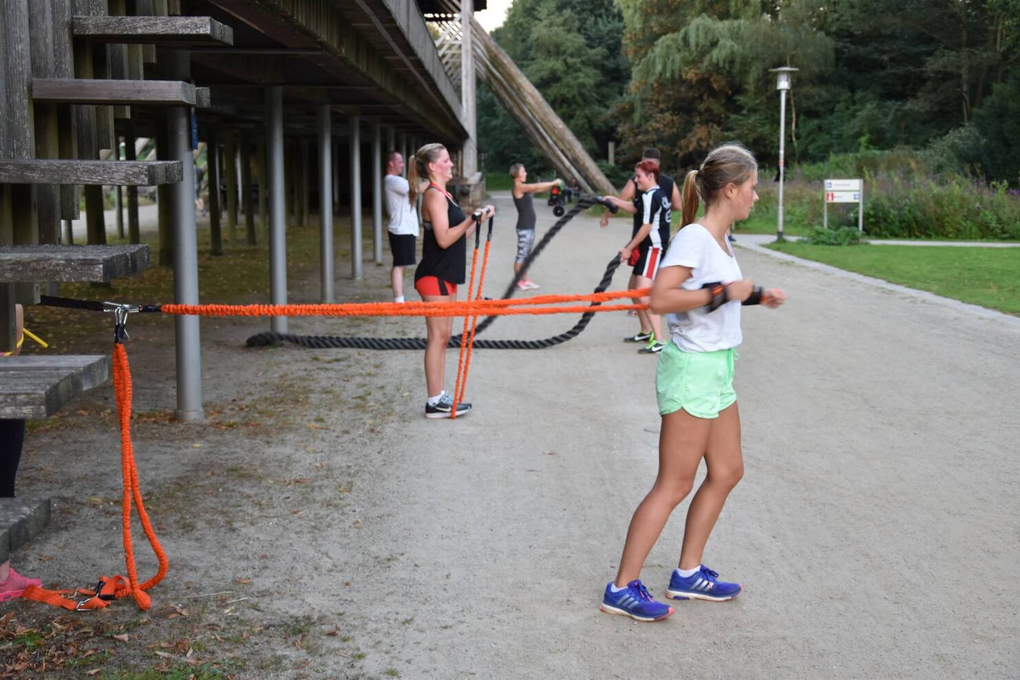 Fitnessgruppe trainiert outdoors mit Widerstandsb&auml;ndern und Reifen auf einem Schotterweg in einer gr&uuml;nen Umgebung.