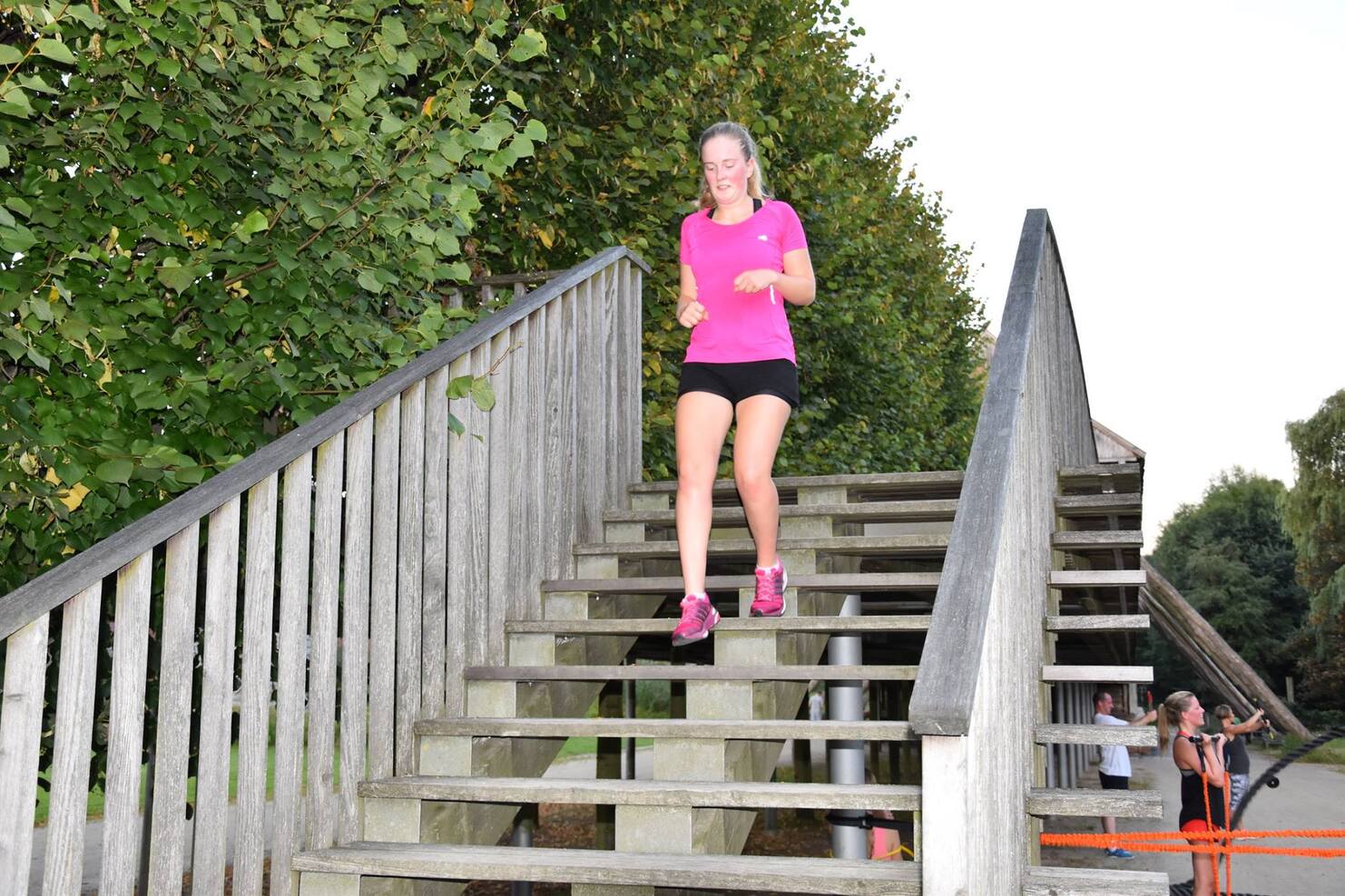 L&auml;uferin in pinkem Shirt l&auml;uft die Holzstufen einer Au&szlig;entreppe hinunter, umgeben von B&auml;umen und anderen Personen im Hintergrund.